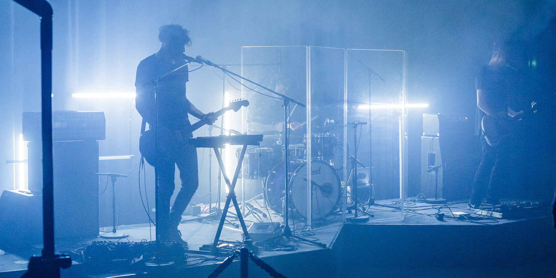 Eau Rouge singer and guitarist silhouetted in blue backlight and haze, drums behind an acrylic screen