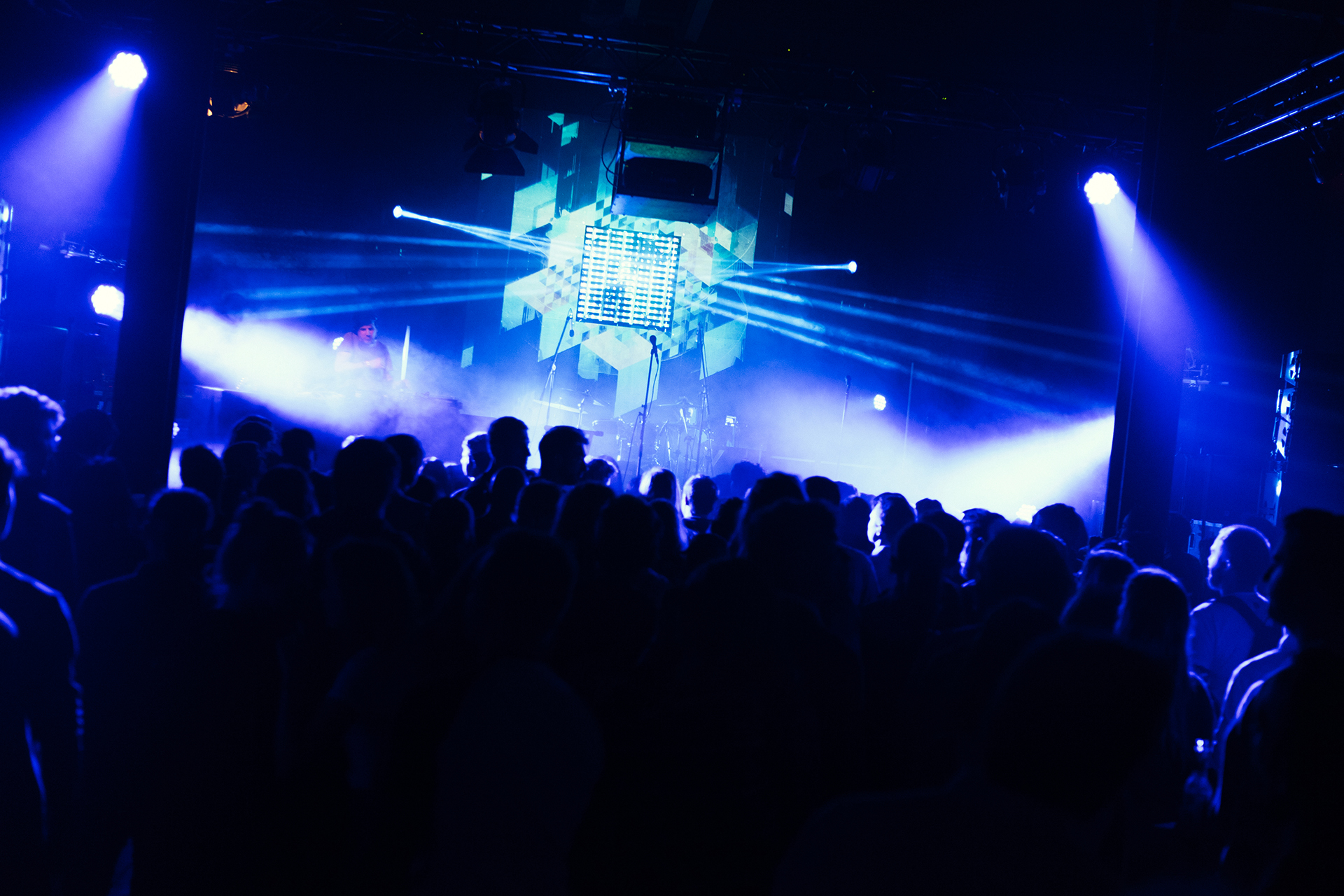 Crowd silhouetted against blue light beams and a geometric projection on the MLIVE stage