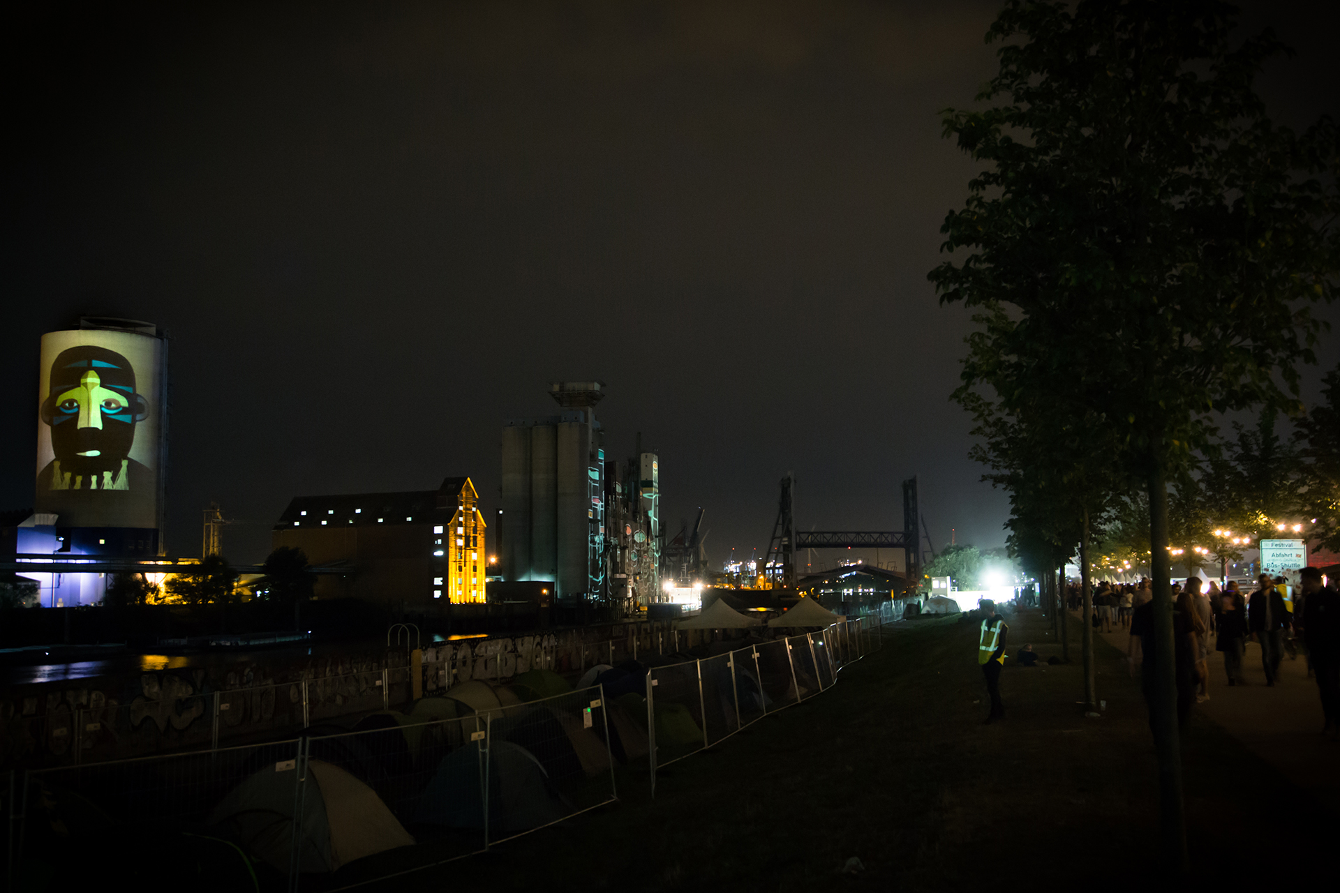 Comic-Porträt des Musikers SBTRKT mit seiner typischen Maske, projiziert auf das Silo über dem Zeltplatz des MS Dockville bei Nacht