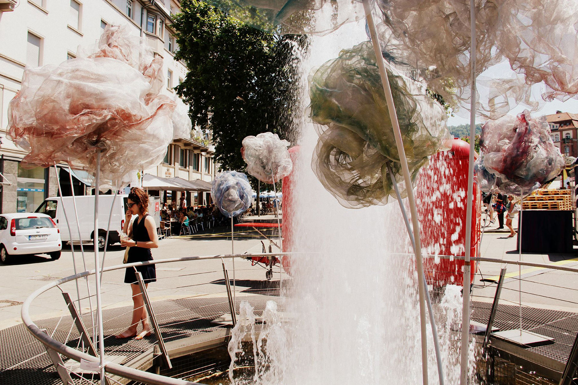 Besucherin geht am Brunnen zwischen durchscheinenden Überblume-Blüten auf dem Marienplatz vorbei